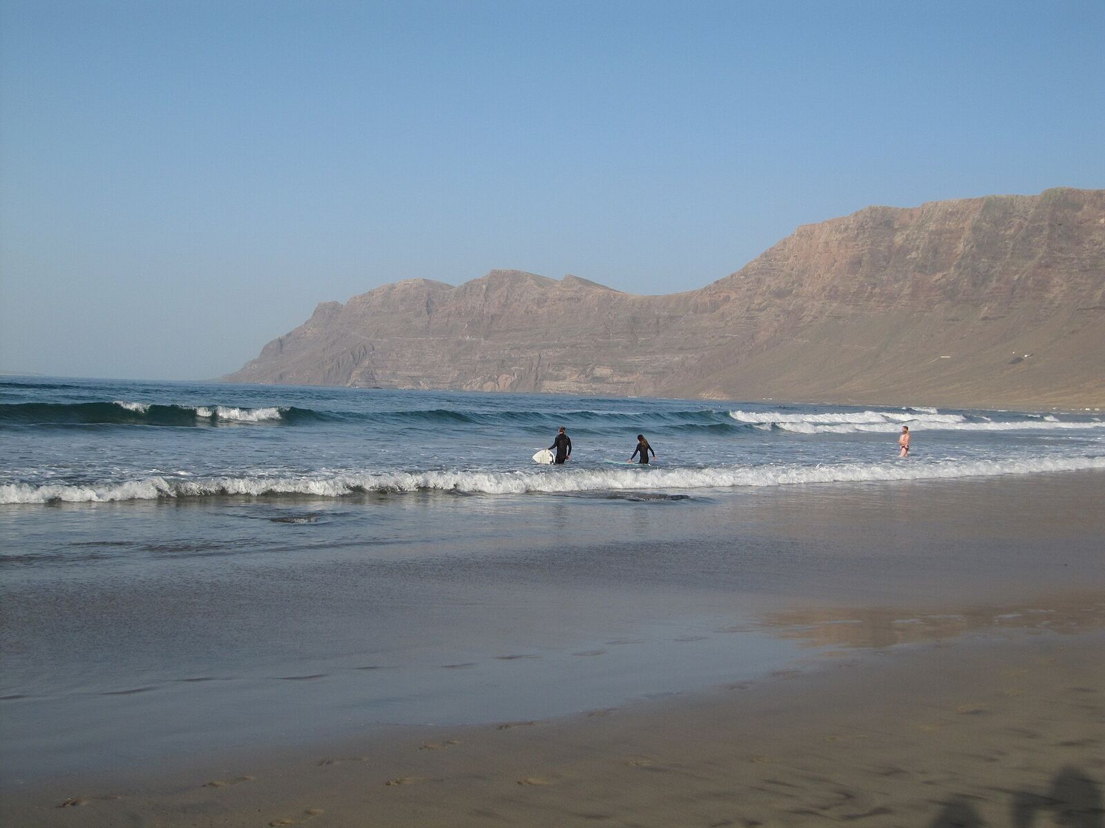 Surfers walking into small waves at Caleta de Famara with the Famara cliffs behind